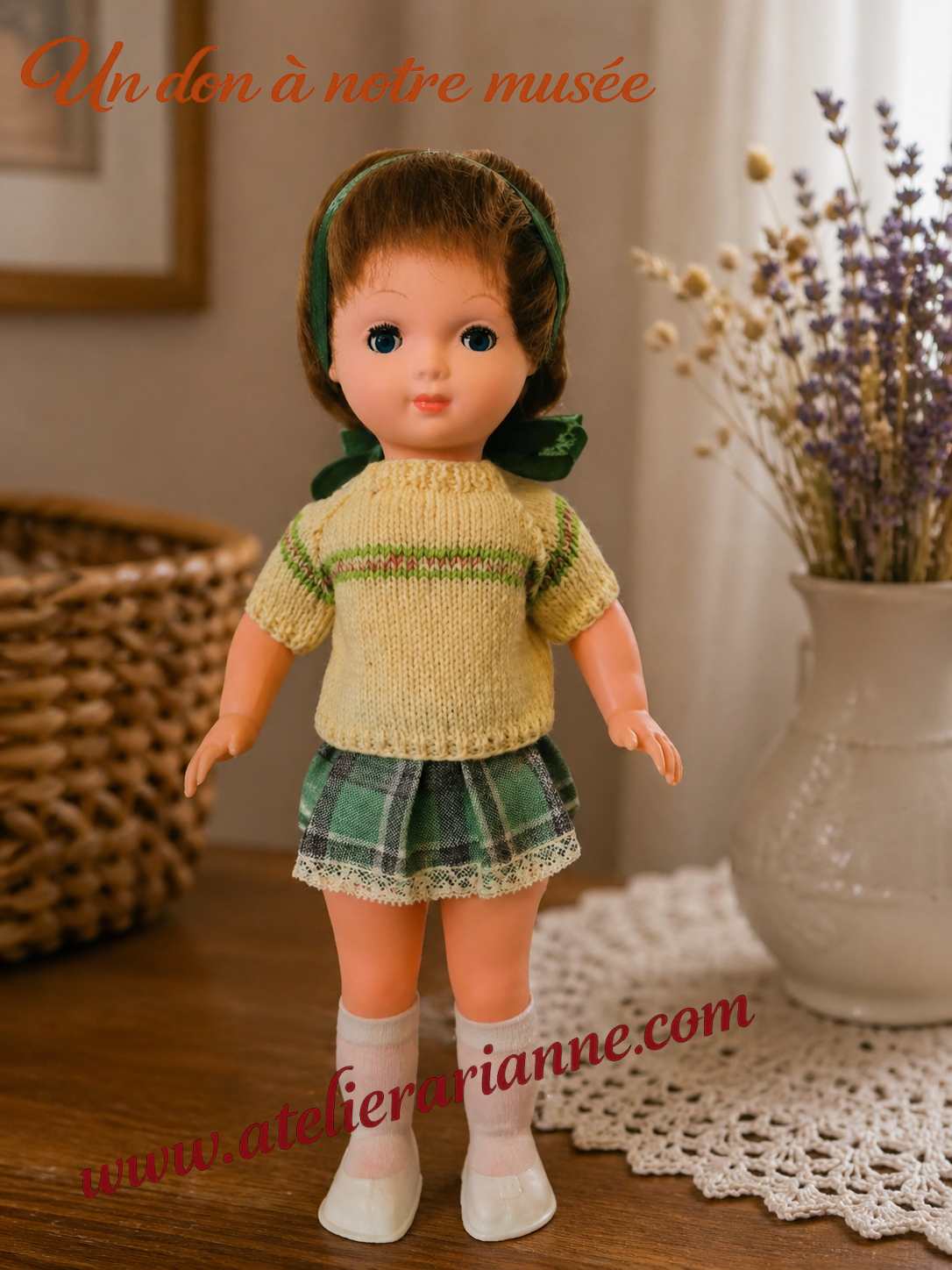 Petite poup&eacute;e Bella restaur&eacute;e pour le mus&eacute;e Atelier Arianne, habill&eacute;e d&rsquo;un pull tricot&eacute;, d&rsquo;une jupe verte, de chaussettes et de chaussures blanches, avec la mention &ldquo;Un don &agrave; notre mus&eacute;e&rdquo;.
